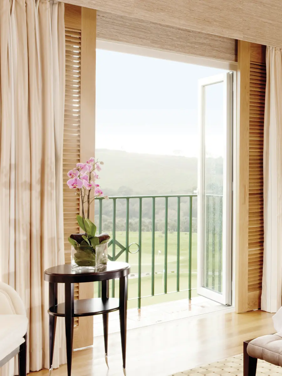 Puerta-ventana de PVC blanco abierta hacia un balcón, con la cortina recogida y vista al campo