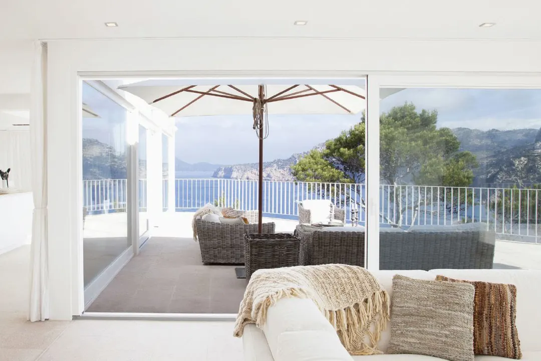Sala abierta a una terraza con sombrilla y vista al mar a través de puertas corredizas de gran claro